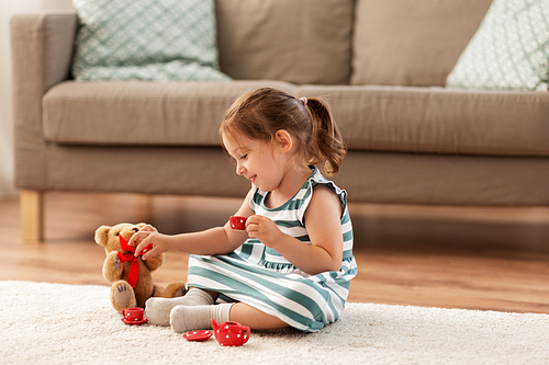 childhood and people concept - happy three years old baby girl playing tea party with toy crockery and teddy bear at home