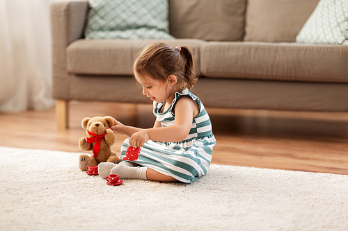 childhood and people concept - happy three years old baby girl playing tea party with toy crockery and teddy bear at home