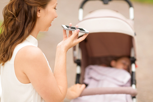 motherhood, technology and people concept - close up of happy mother with baby in stroller using voice command recorder on smartphone at summer park