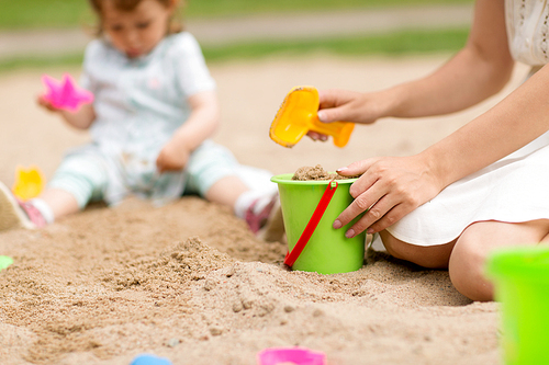 childhood, leisure and people concept - close up of mother with sand in toy bucket playing with little baby daughter in sandbox