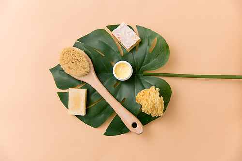 beauty, spa and bath concept - close up of crafted soap bar, natural bristle wooden brush, body butter with sponge and monstera deliciosa leaf on beige background
