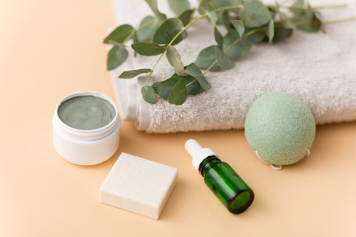 beauty, spa and wellness concept - serum or essential oil, clay mask, soap bar, konjac sponge and eucalyptus cinerea on bath towel