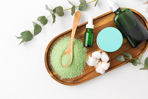beauty and spa concept - green bath salt, serum with dropper, body oil, moisturizer and eucalyptus cinerea with cotton flowers on wooden tray