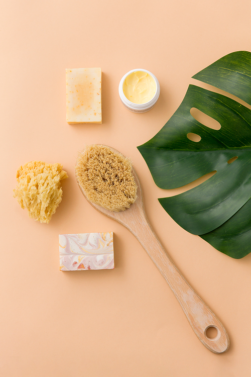 beauty, spa and bath concept - close up of crafted soap bar, natural bristle wooden brush, body butter with sponge and monstera deliciosa leaf on beige background
