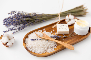 beauty, bath and wellness concept - sea salt with spoon, soap bar, body butter, lavender and cotton flowers on wooden tray