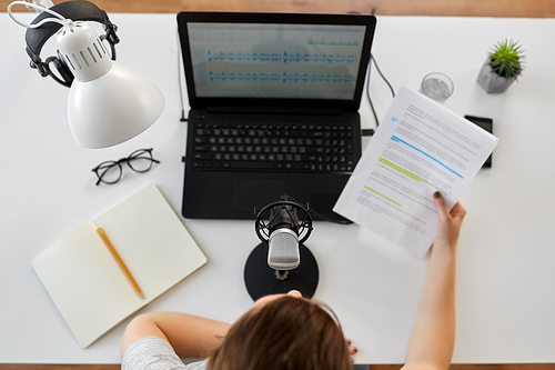 technology, mass media and people concept - woman with microphone and laptop computer reading text from papers and recording podcast at studio