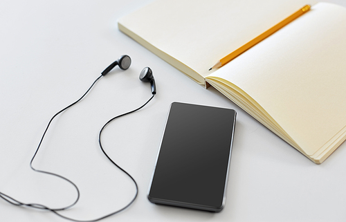 technology and objects concept - earphones, smartphone and notebook with pencil on white background