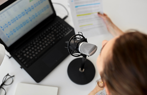 technology, mass media and people concept - woman with microphone and laptop computer reading text from papers and recording podcast at studio