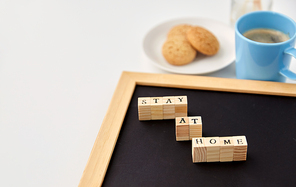 quarantine, epidemic and safety concept - close up of chalkboard with stay at home words on wooden toy blocks, coffee cup, cookies and aroma reed diffuser on white background
