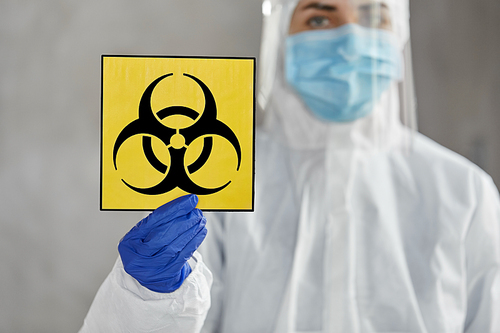 health safety, medicine and pandemic concept - close up of female doctor or scientist in protective wear, medical mask and face shield for protection from virus disease holding biohazard caution sign