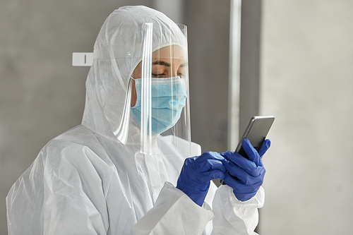 health safety, medicine and pandemic concept - close up of female doctor or scientist in protective wear, medical mask, gloves and face shield for protection from virus disease with smartphone