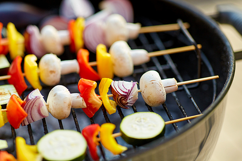 cooking, barbecue and food concept - close up of vegetables with champignon mushrooms on bamboo skewers roasting on hot brazier grill outdoors