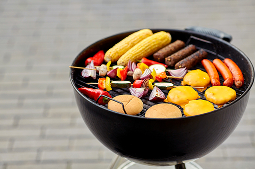 cooking, barbecue and food concept - close up of shish kebab meat, vegetables on bamboo skewers, burger buns with corn and sausages roasting on brazier grill outdoors