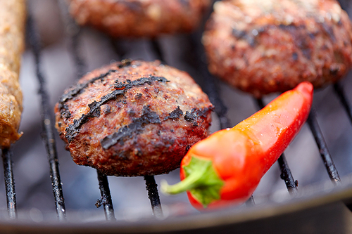 cooking, barbecue and food concept - close up of burger meat cutlets and red chili pepper roasting on brazier grill outdoors