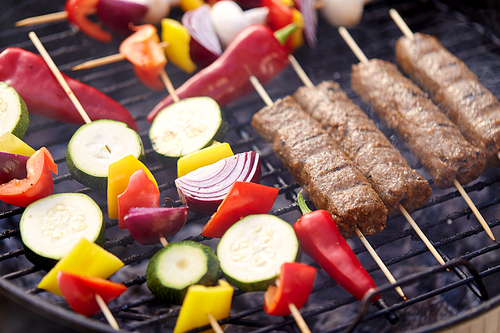 cooking, barbecue and food concept - close up of shish kebab meat and vegetables on bamboo skewers roasting to hot brazier grill outdoors
