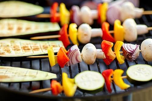 cooking, barbecue and food concept - close up of eggplant slices and vegetables with champignon mushrooms on bamboo skewers roasting on hot brazier grill outdoors