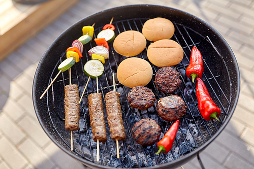 cooking, barbecue and food concept - close up of shish kebab meat, vegetables on bamboo skewers, burger buns and cutlets roasting on brazier grill outdoors
