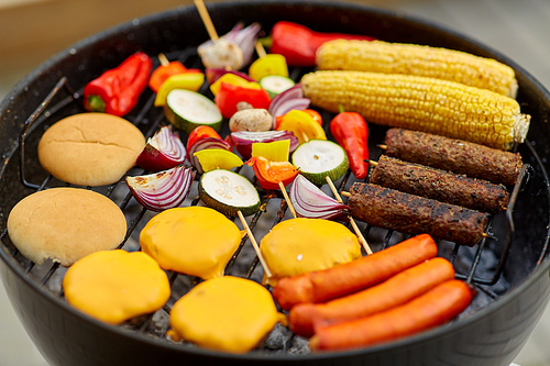 cooking, barbecue and food concept - close up of shish kebab meat, vegetables on bamboo skewers, burger buns with corn and sausages roasting on brazier grill outdoors