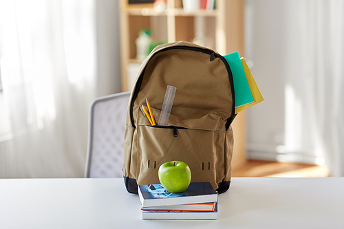 education and learning concept - backpack with books, school supplies and green apple on table at home