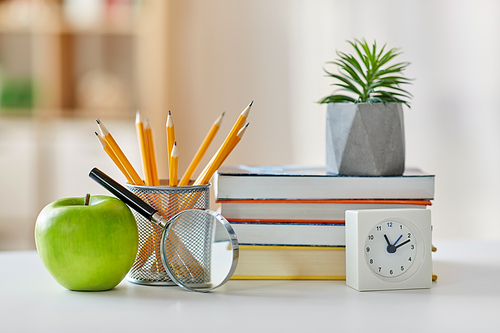 education and school concept - books, magnifier, pencils, plant in flower pot with green apple and alarm clock on table at home