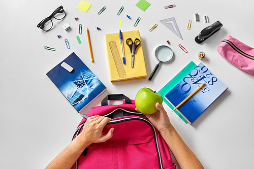 education and learning concept - hands packing pink backpack with green apple, books and school supplies on table