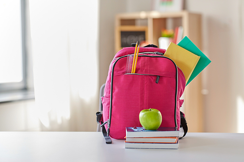 education and learning concept - pink backpack with books and school supplies, green apple on table at home