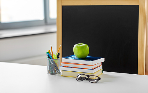 education and learning concept - books, apple, chalkboard and school supplies on table at home