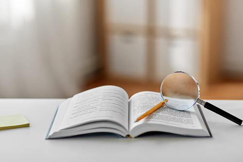 education, school and learning concept - book with magnifier and pencil on table at home