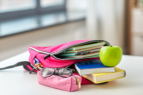 education and learning concept - close up of pink backpack with books and school supplies, green apple on table at home