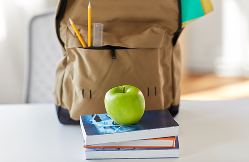 education and learning concept - close up of backpack with books, school supplies and green apple on table at home