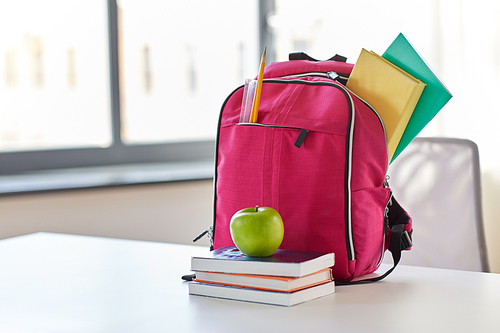 education and learning concept - pink backpack with books and school supplies, green apple on table at home