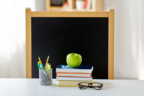 education and learning concept - books, apple, chalkboard and school supplies on table at home