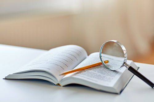 education, school and learning concept - book with magnifier and pencil on table at home