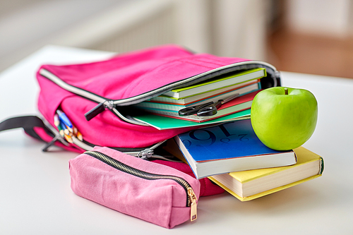 education and learning concept - close up of pink backpack with books and school supplies, green apple on table at home