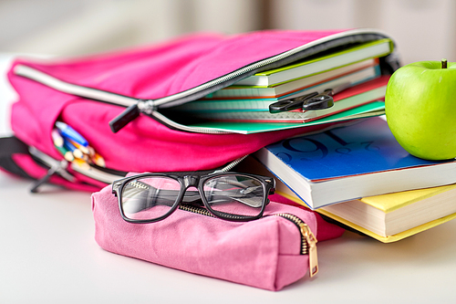 education and learning concept - close up of pink backpack with books and school supplies, green apple on table at home