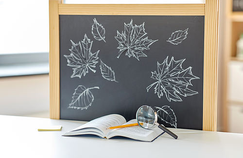 education and learning concept - book, magnifier and pencil on table and autumn leaves drawn on chalkboard at home