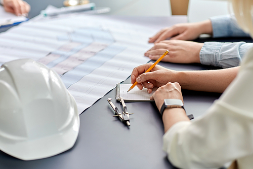 construction business, architecture and buiding concept - close up of architect with blueprint, notebook and pencil working at office