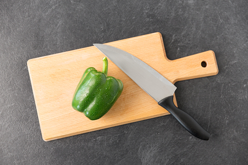 vegetable, food and culinary concept - close up of green pepper and kitchen knife on wooden cutting board on slate stone background