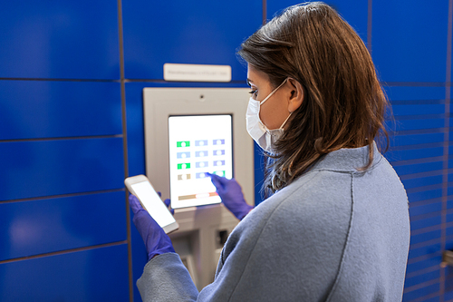 health care, mail delivery and pandemic concept - woman wearing face protective medical mask for protection from virus disease with smartphone entering code on automated parcel machine's touch screen