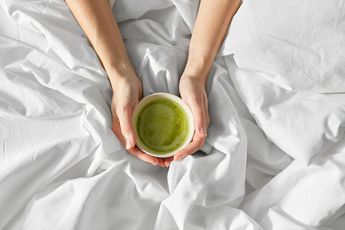 morning, drink and people concept - hands of young woman with cup of matcha tea in bed at home bedroom