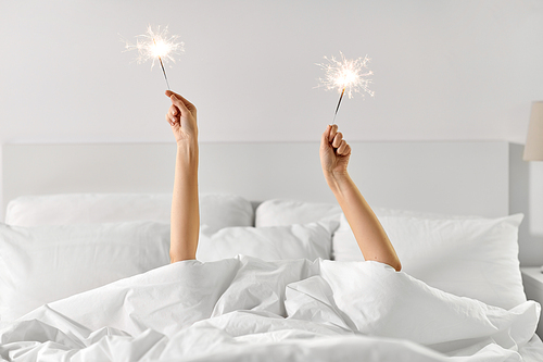 celebration, comfort and morning concept - hands of young woman lying in bed with sparklers