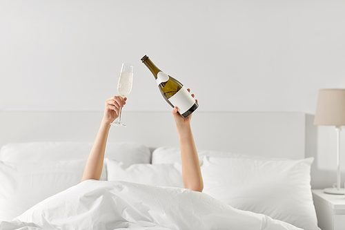 alcohol, comfort and morning concept - hands of young woman lying in bed with champagne glass and bottle