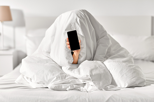 technology, comfort and morning concept - hand of young woman sitting in bed under blanket with smartphone at bedroom