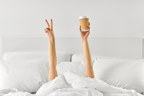 morning, comfort and people concept - young woman with cup of takeaway coffee lying in bed and showing peace hand sign at home bedroom