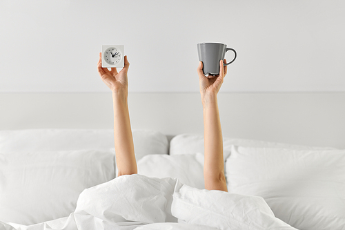 morning, comfort and people concept - young woman with cup of coffee and alarm clock lying in bed at home bedroom