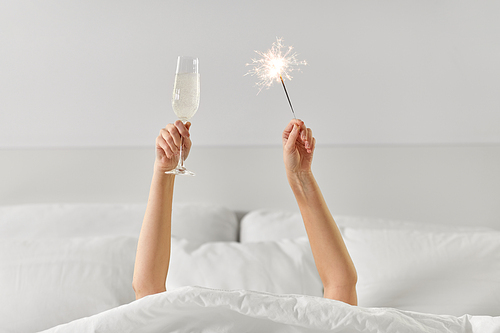 alcohol, celebration and morning concept - hands of young woman lying in bed with champagne glass and sparkler