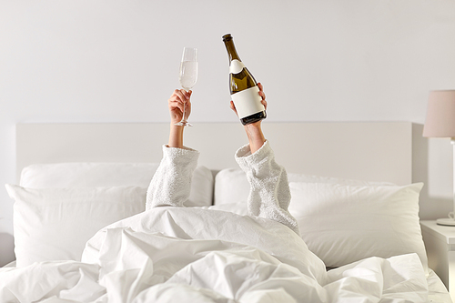 alcohol, celebration and morning concept - hands of young woman in hotel robe lying in bed with champagne glass and bottle