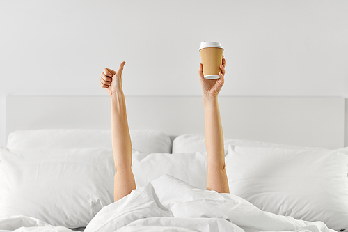 morning, comfort and people concept - young woman with cup of takeaway coffee lying in bed and showing thumbs up at home bedroom