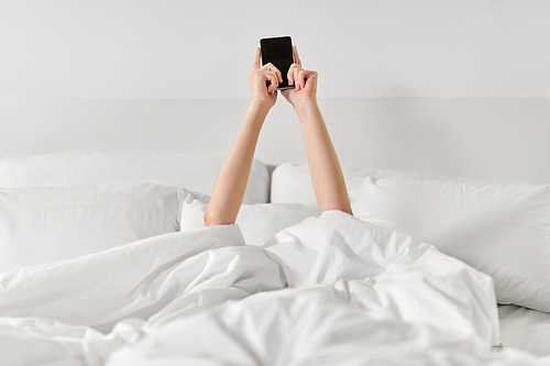 technology, comfort and morning concept - hands of young woman lying in bed with smartphone at bedroom