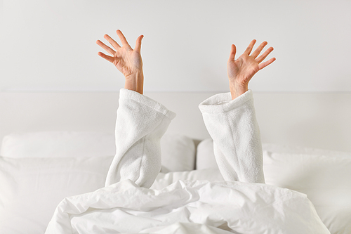 gesture, comfort and morning concept - hands of young woman in hotel robe lying in bed and stretching at bedroom